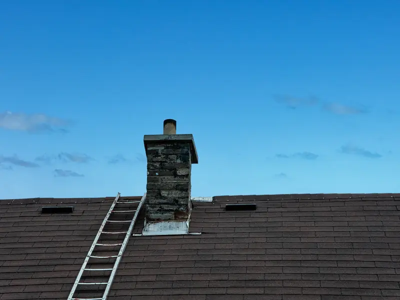 Brick chimney with metal flashing at its base on a shingle roof, a ladder laid up the slope