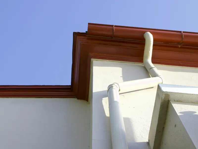Brown gutter wrapping a roof corner with white downspouts against a blue sky