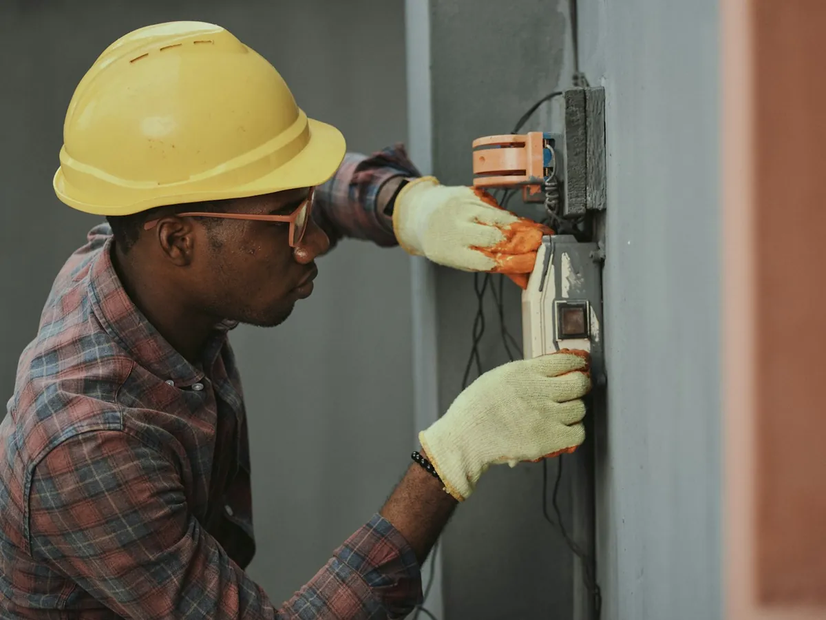 An electrician in a hard hat and work gloves wiring a box mounted on a wall