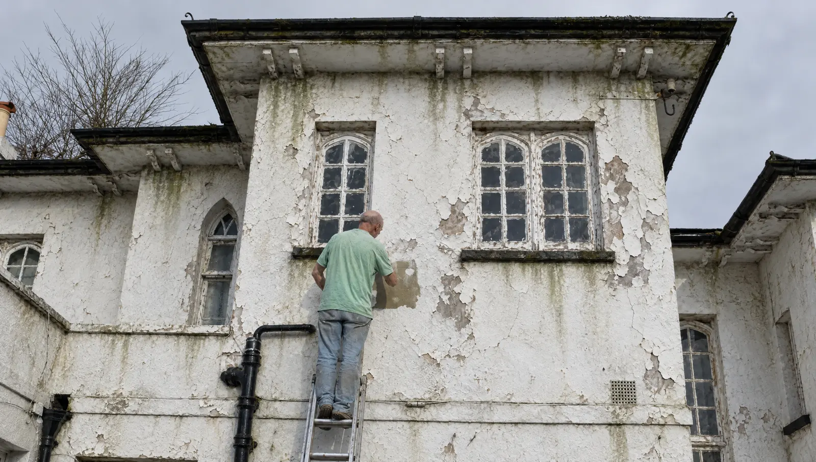 A painter on a ladder at a house with peeling, stained white walls